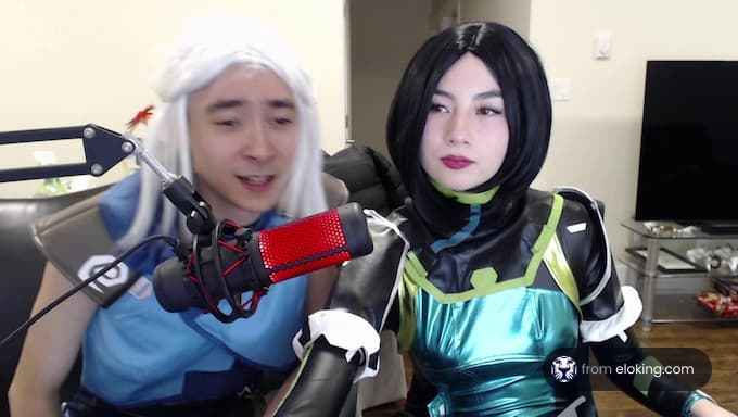 Two people in cosplay as characters from a video game, one dressed in white and blue with a wig, the other in a green and black suit, sitting with a microphone