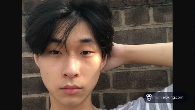 A young man with black hair posing against a brick wall on a sunny day.